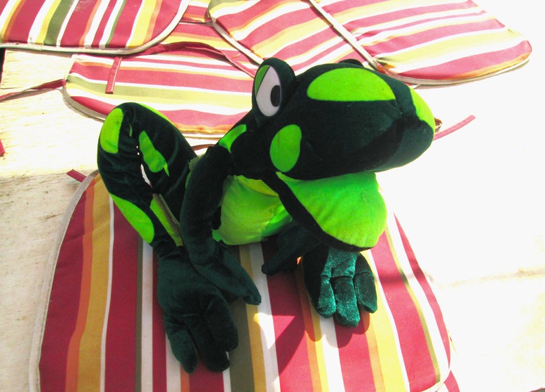 Pu&ograve; includere: Un giocattolo di peluche di rana verde e nero con grandi occhi rotondi. La rana &egrave; seduta su un cuscino a strisce rosso, giallo e bianco.