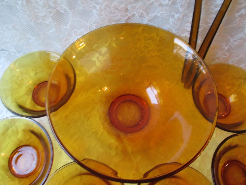 Peut inclure: Ensemble de bols de service en verre ambr&eacute;. Les bols sont ronds avec une base centrale sur&eacute;lev&eacute;e et un bord large et peu profond. Le verre a une finition brillante, refl&eacute;tant la lumi&egrave;re. Les bols sont dispos&eacute;s sur une nappe en dentelle blanche.