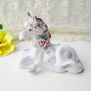 May include: A white porcelain unicorn figurine with a pink and white floral crown, lying down with its head turned to the side. The unicorn has a grey mane and tail.