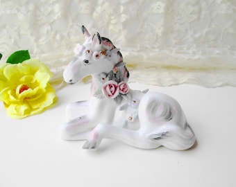 Puede incluir: Una figurita de unicornio de porcelana blanca con una corona floral rosa y blanca, tumbada con la cabeza girada hacia un lado. El unicornio tiene una melena y una cola grises.