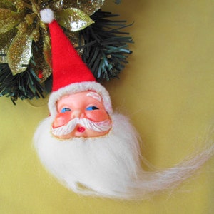 May include: A vintage Santa Claus ornament with a red hat, white beard, and a plastic face. The ornament is hanging from a gold and green garland.