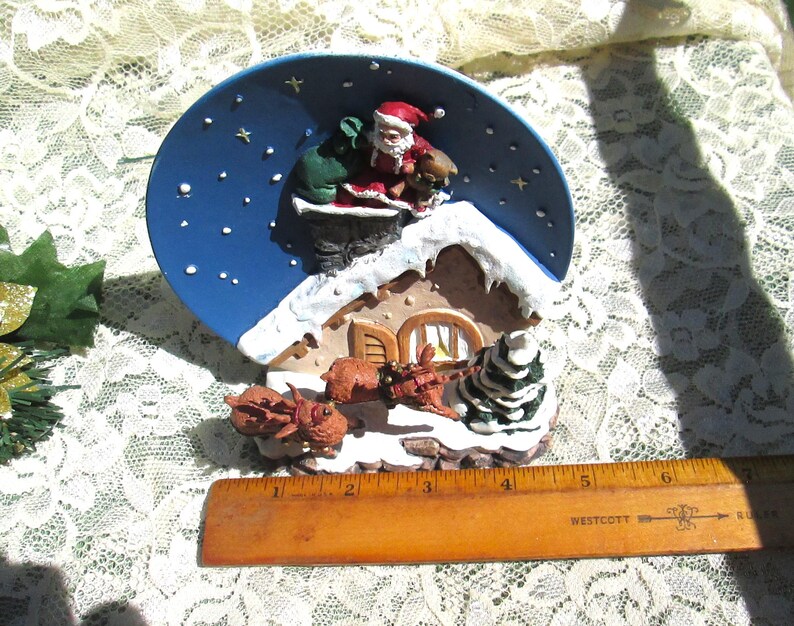 Może przedstawiać: Ceramiczna figurka świąteczna przedstawiająca Świętego Mikołaja schodzącego po kominku z workiem prezent&oacute;w. Figurka stoi na niebiesko-białym talerzu z ośnieżonym dachem i choinką. Dwa renifery ciągną sanie przed domem.