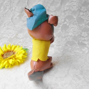 Puede incluir: Un juguete de peluche marr&oacute;n con un sombrero azul y una camisa amarilla. El juguete est&aacute; de pie sobre un fondo de encaje blanco.