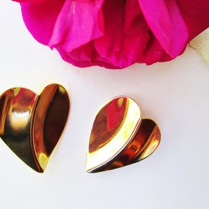 May include: A pair of gold-tone heart-shaped earrings. The earrings are made of two overlapping pieces of metal, creating a layered effect.