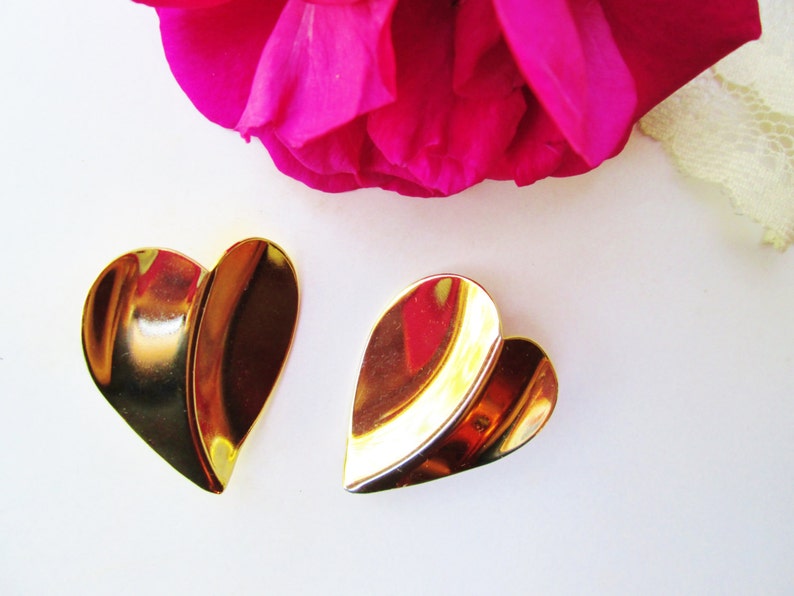 May include: A pair of gold-tone heart-shaped earrings. The earrings are made of two overlapping pieces of metal, creating a layered effect.