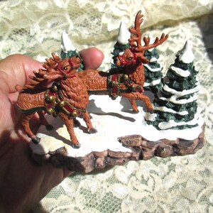 Może przedstawiać: Miniaturowa scena świąteczna z dwoma brązowymi reniferami ciągnącymi sanie, otoczonymi trzema małymi, ośnieżonymi choinkami. Renifery są ozdobione czerwonymi i złotymi akcentami.