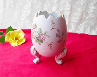 May include: White ceramic egg-shaped vase with a cracked top and gold trim. The vase is decorated with pink and green roses and has three gold-trimmed feet.