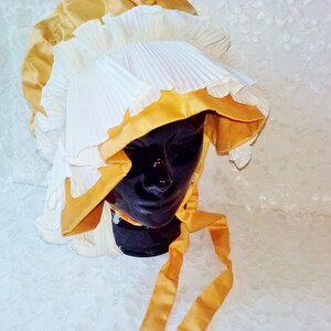 Puede incluir: Un gorro blanco y dorado con un dise&ntilde;o plisado. El gorro tiene un ala ancha y est&aacute; adornado con una cinta dorada.