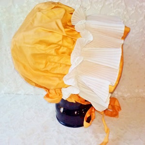 Puede incluir: Un gorro de sat&eacute;n amarillo con ribete de encaje blanco. El gorro est&aacute; atado con una cinta amarilla.