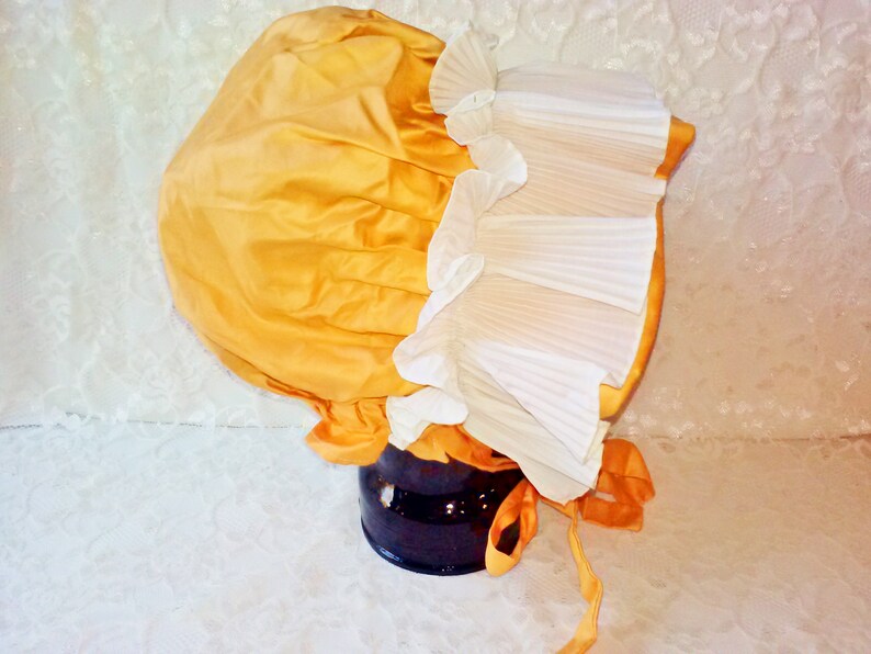 Puede incluir: Un gorro de sat&eacute;n amarillo con ribete de encaje blanco. El gorro est&aacute; atado con una cinta amarilla.