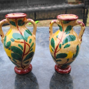 May include: Two small ceramic vases with handles, painted with a yellow, green, and red floral design.