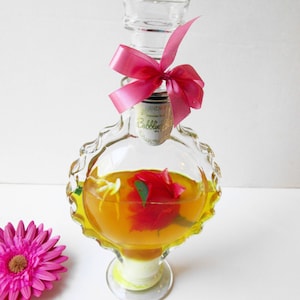 May include: A clear glass bottle with a pink ribbon and a single red rose inside. The bottle is filled with a yellow liquid and has a label that reads "Bubbles Oil".