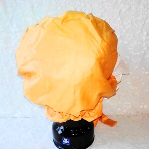 Puede incluir: Un gorro de ducha de tela de color naranja brillante con un ribete de encaje blanco. El gorro est&aacute; sobre una superficie blanca.