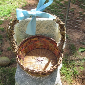 May include: A woven wicker basket with a light blue ribbon tied around the handle. The basket is empty and sitting on a white lace tablecloth.