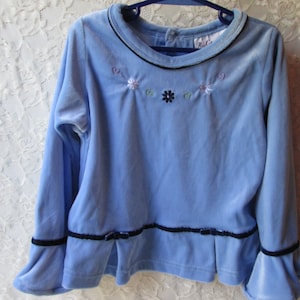 May include: A light blue long-sleeved shirt with a black trim around the neckline and cuffs. The shirt has a small embroidered design of flowers on the front.