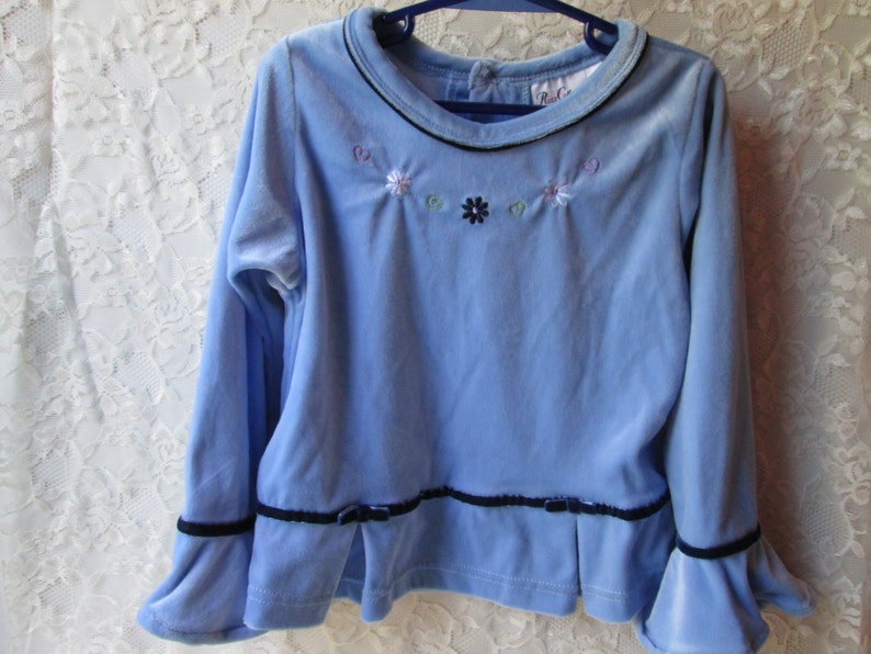 May include: A light blue long-sleeved shirt with a black trim around the neckline and cuffs. The shirt has a small embroidered design of flowers on the front.