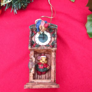May include: A Christmas ornament shaped like a grandfather clock. The clock face is white with black numbers and hands. A small mouse wearing a Santa hat and a red and green outfit is inside the clock. The ornament is brown and has a gold string for hanging.