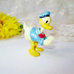 May include: A yellow plastic Donald Duck figurine wearing a blue sailor hat and a red bow tie. The figurine is standing on a white surface.