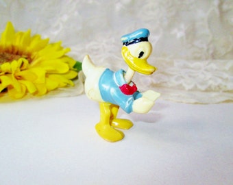Puede incluir: Una figura de plástico amarillo de Donald Duck con un sombrero de marinero azul y una pajarita roja. La figura está de pie sobre una superficie blanca.