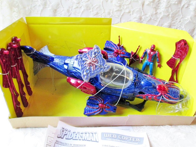 May include: A blue and red plastic toy helicopter with a spider web design. The helicopter is in a yellow box with a Spider-Man figure and instructions. The box says "The Amazing Spider-Man Web Copter".