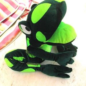 Pu&ograve; includere: Un giocattolo di peluche a forma di rana verde con macchie nere. La rana &egrave; seduta su una superficie bianca con le gambe incrociate.