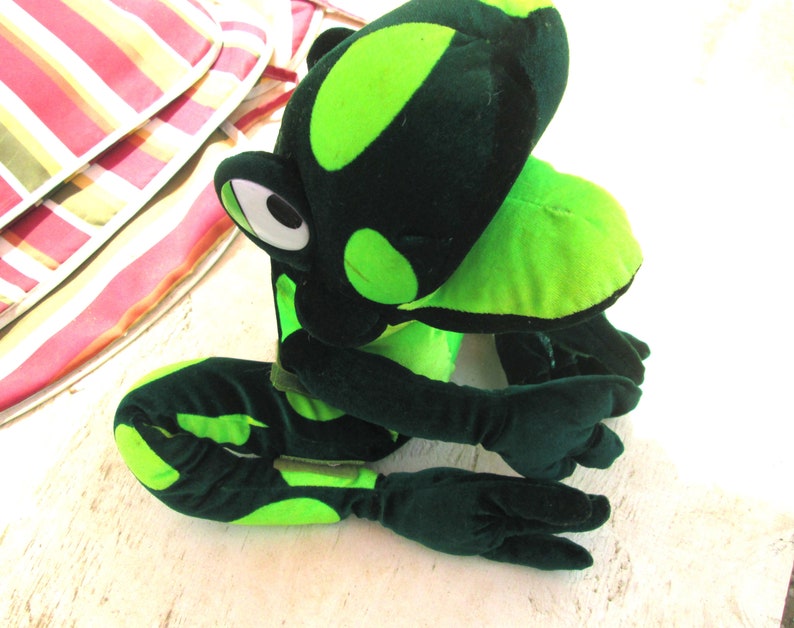 Pu&ograve; includere: Un giocattolo di peluche a forma di rana verde con macchie nere. La rana &egrave; seduta su una superficie bianca con le gambe incrociate.