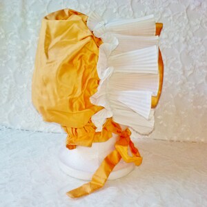 Puede incluir: Un gorro de sat&eacute;n amarillo con un volante de encaje blanco. El gorro est&aacute; atado con una cinta de sat&eacute;n amarilla.