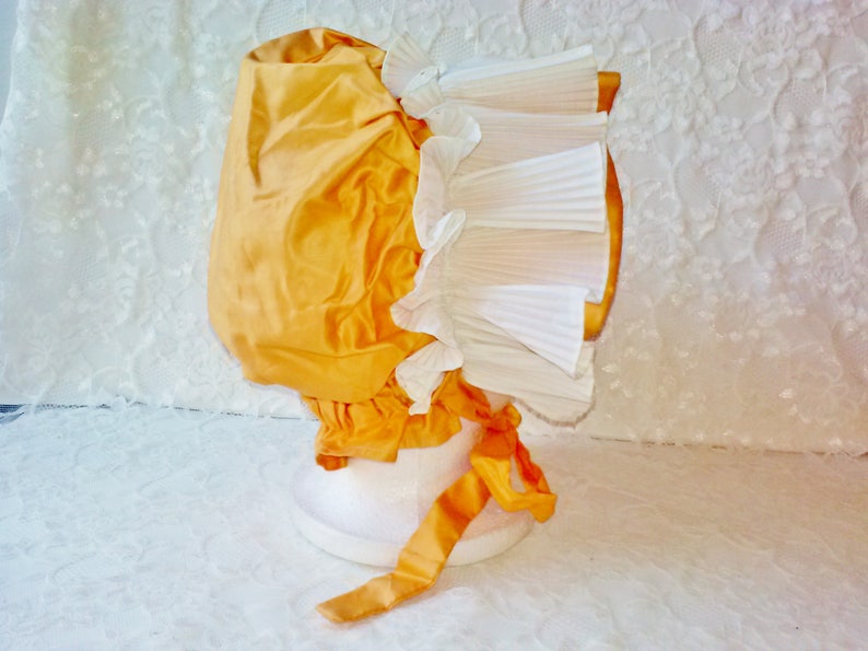 Puede incluir: Un gorro de sat&eacute;n amarillo con un volante de encaje blanco. El gorro est&aacute; atado con una cinta de sat&eacute;n amarilla.