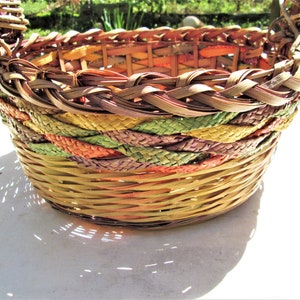 May include: A woven basket with a colorful design. The basket is made of natural materials and has a handle. The basket is empty and sitting on a white surface.