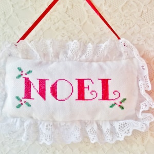 May include: White fabric pillow with a red embroidered "NOEL" and green holly sprigs. The pillow is trimmed with white lace and has a red ribbon for hanging.