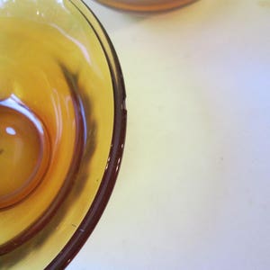 Peut inclure: Gros plan sur un bol en verre ambr&eacute; vintage avec une indentation circulaire au centre. Le bol a une surface lisse et brillante et un bord ambr&eacute; fonc&eacute;. Le bol est pos&eacute; sur un fond blanc.