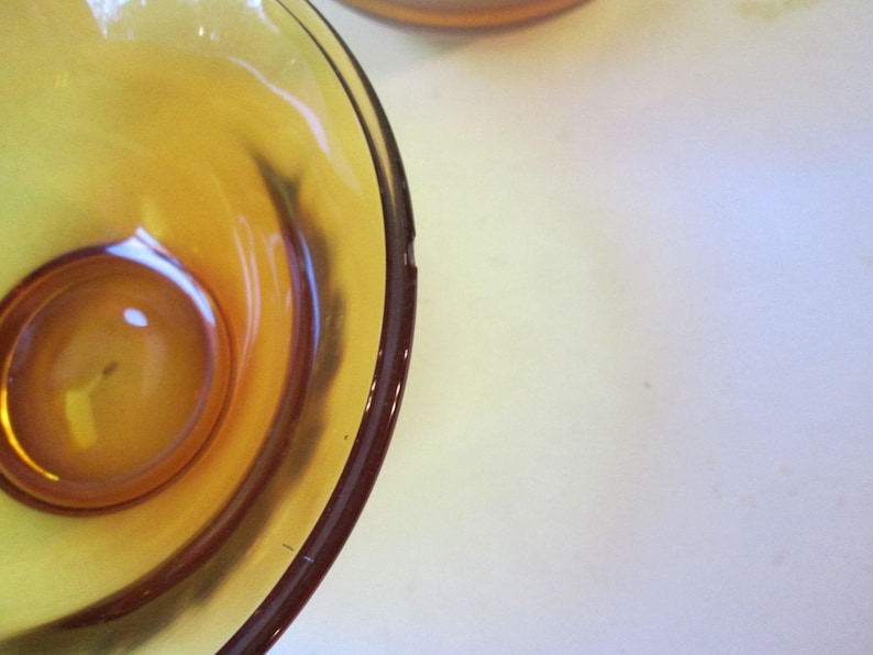 Peut inclure: Gros plan sur un bol en verre ambr&eacute; vintage avec une indentation circulaire au centre. Le bol a une surface lisse et brillante et un bord ambr&eacute; fonc&eacute;. Le bol est pos&eacute; sur un fond blanc.