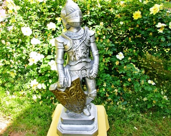 May include: A silver knight statue with a shield and sword, standing on a pedestal in front of a green bush with white flowers.