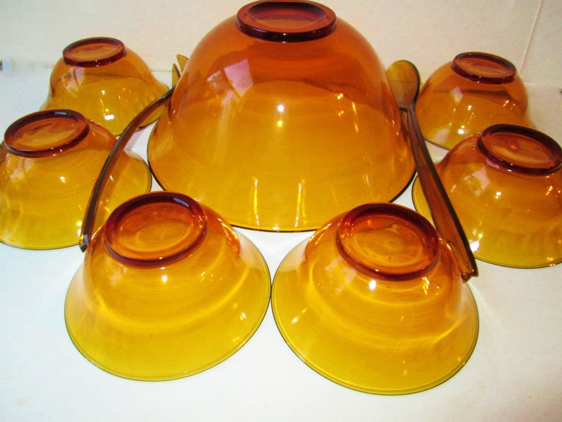 Peut inclure: Un ensemble de service en verre ambr&eacute; vintage, comprenant un grand saladier, six petits bols et deux cuill&egrave;res de service. Les bols et les cuill&egrave;res ont une teinte ambr&eacute;e chaude et transparente.