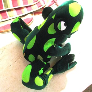 Pu&ograve; includere: Un giocattolo di peluche di rana verde e nero con grandi occhi bianchi. La rana ha un motivo a pois ed &egrave; seduta su una superficie bianca.