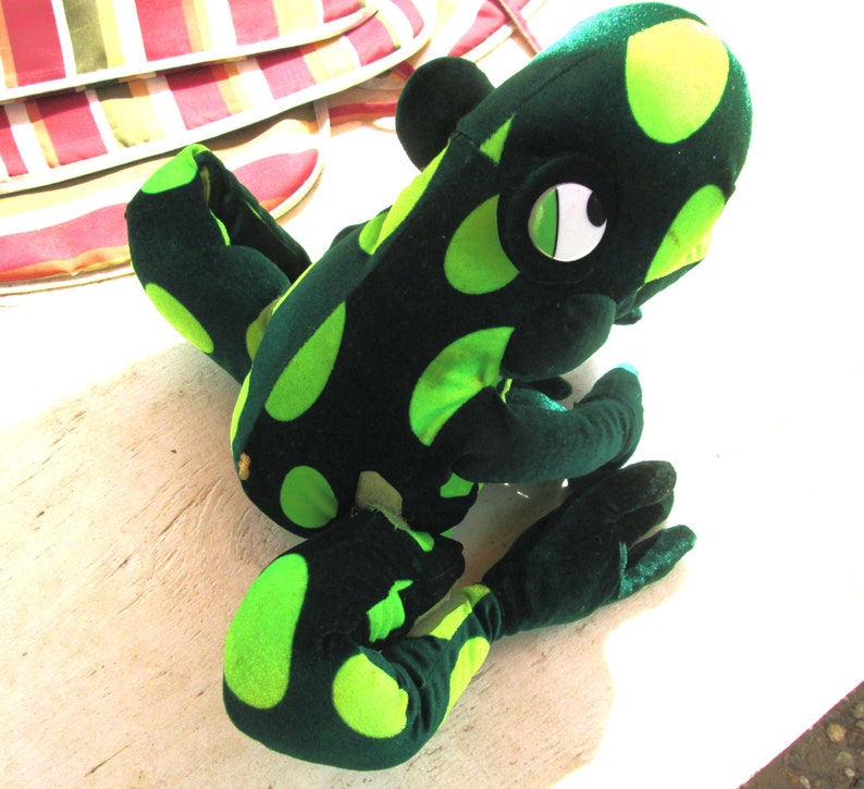 Pu&ograve; includere: Un giocattolo di peluche di rana verde e nero con grandi occhi bianchi. La rana ha un motivo a pois ed &egrave; seduta su una superficie bianca.