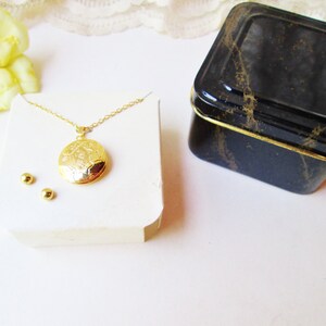 May include: A gold locket necklace with a detailed floral design on the front. The necklace is displayed on a white card with two small gold earrings. The necklace is packaged in a black and gold tin box.