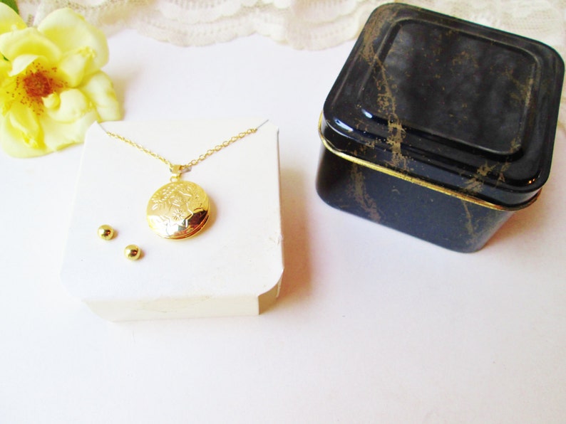 May include: A gold locket necklace with a detailed floral design on the front. The necklace is displayed on a white card with two small gold earrings. The necklace is packaged in a black and gold tin box.