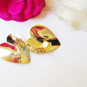 May include: A pair of gold heart-shaped earrings with a clip-on closure. The earrings are shiny and have a slightly textured surface.