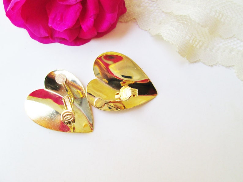 May include: A pair of gold heart-shaped earrings with a clip-on closure. The earrings are shiny and have a slightly textured surface.