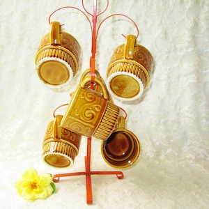May include: Set of five brown ceramic mugs with a decorative swirl pattern hanging on a red metal rack.