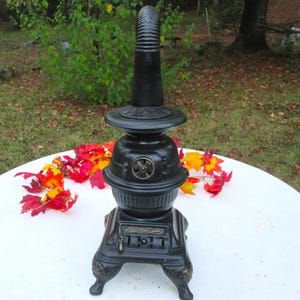 May include: A black cast iron antique stove with a tall, ribbed chimney. The stove has a round body with a small door and decorative details. The stove is sitting on a white surface with colorful fall leaves scattered around it.