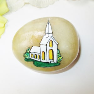 May include: Hand-painted rock with a white church building with a steeple and yellow windows, surrounded by green bushes on a green lawn.