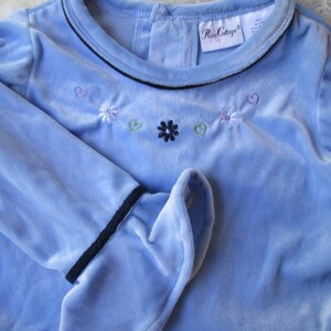 May include: A light blue long-sleeved shirt with a black trim around the neckline and cuffs. The shirt has embroidered flowers and hearts in shades of pink, purple, green, and blue.