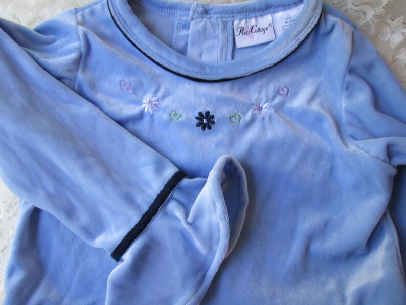 May include: A light blue long-sleeved shirt with a black trim around the neckline and cuffs. The shirt has embroidered flowers and hearts in shades of pink, purple, green, and blue.