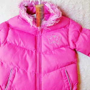 May include: A pink puffer jacket with a white faux fur trim around the hood. The jacket has a zip closure and a butterfly embroidered on the front.