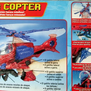 May include: A red and blue toy helicopter with a spider-web design. The helicopter has a clear cockpit and a cannon on top. The box features text in Spanish and Portuguese describing the features of the toy.