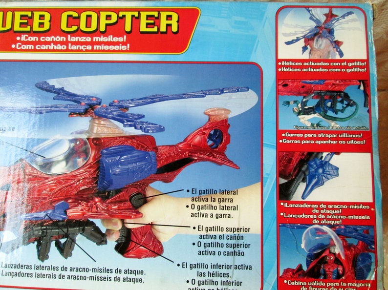 May include: A red and blue toy helicopter with a spider-web design. The helicopter has a clear cockpit and a cannon on top. The box features text in Spanish and Portuguese describing the features of the toy.