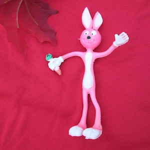 May include: A pink, bendable bunny toy with white gloves and feet, holding a carrot. The bunny has long ears and is posed with one arm raised. The background is a solid red color.