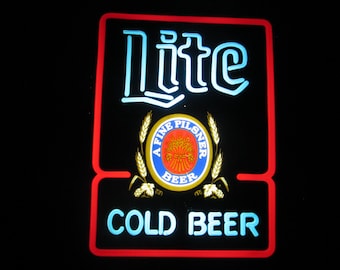 Puede incluir: Un letrero de neón rojo, blanco y azul con las palabras "Lite" y "Cold Beer". El letrero también presenta un logotipo circular con las palabras "A Fine Pilsner Beer".